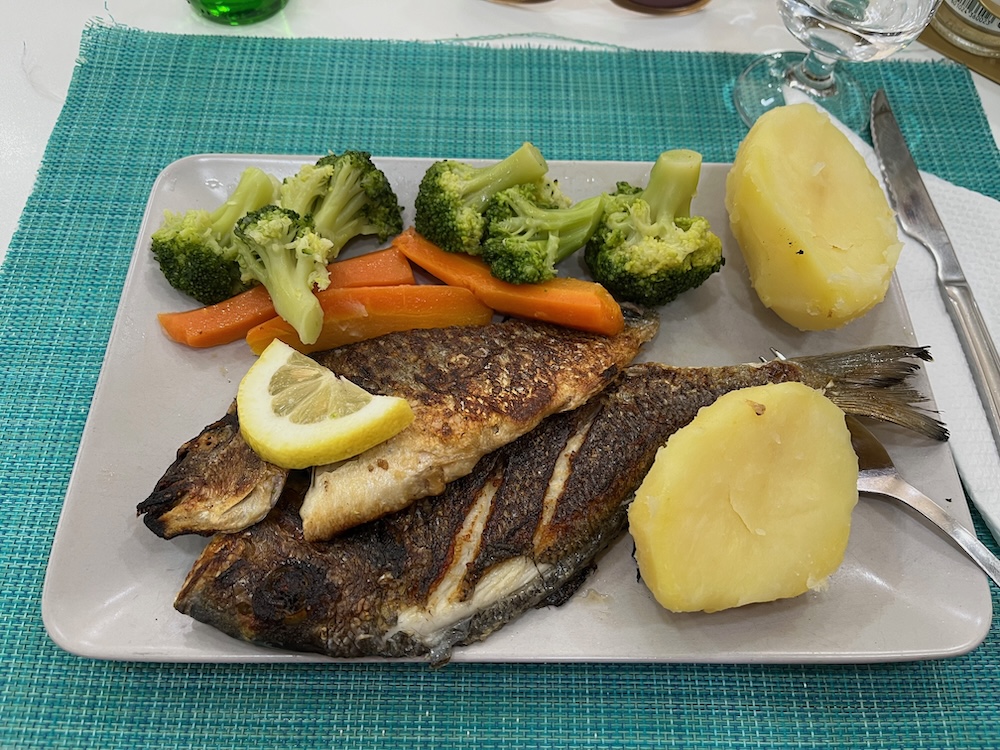 A whole fish (probably dourada) that was cut in half lengthwise and grilled, on a plate with boiled potatoes, broccoli, and carrots, with a slice of lemon on the fish.
